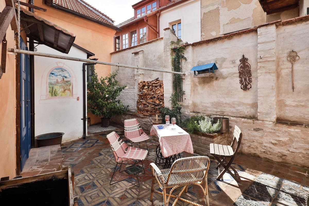A quaint outdoor courtyard features patterned stone flooring. Two patterned chairs and a small table are arranged under the warm sun. A mural adorns the wall, and a stack of firewood is neatly placed nearby, complemented by greenery and a blue decorative element.