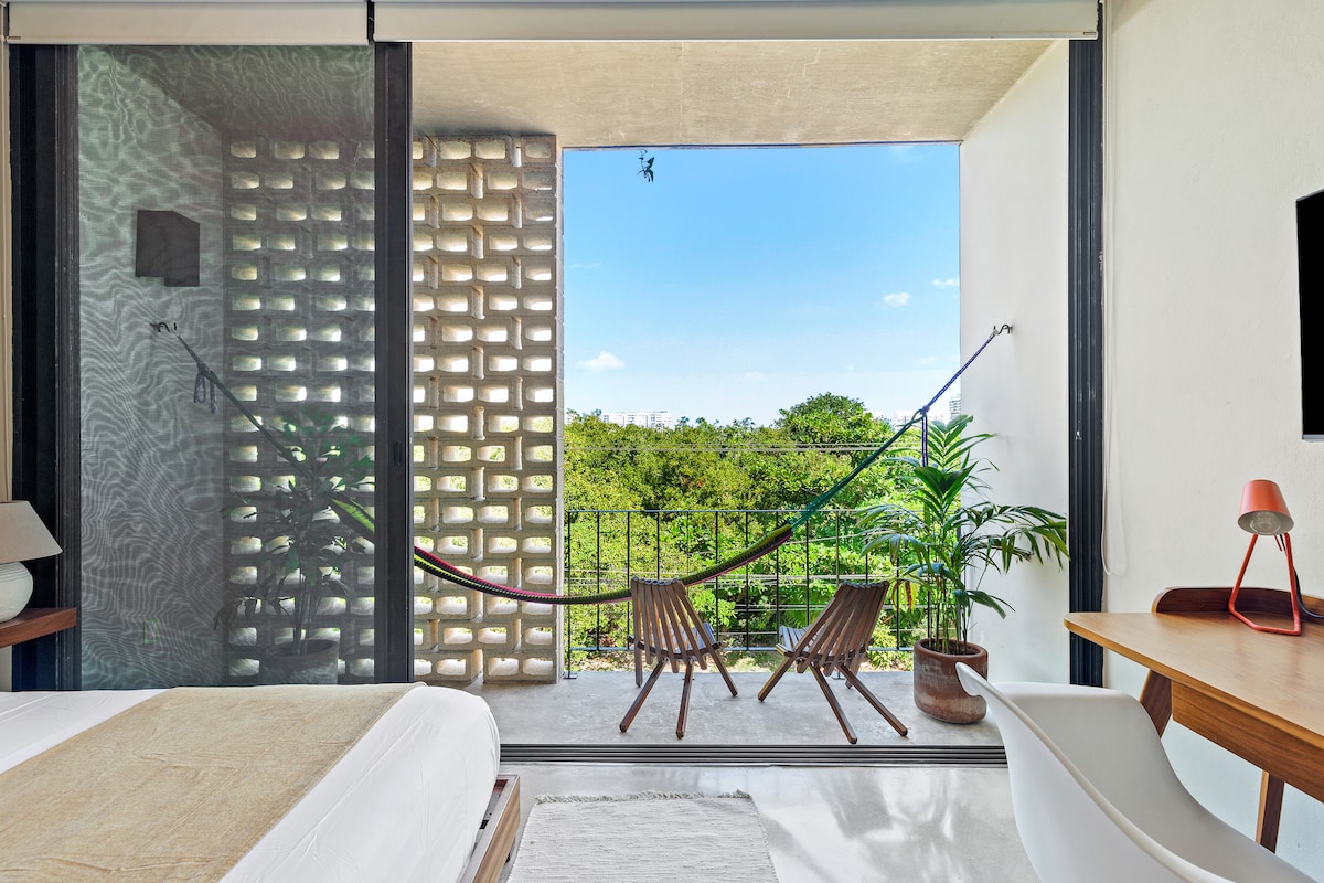 A spacious area features a comfortable queen-sized bed, a desk, and a modern TV. Natural light floods in through large sliding doors that open to a private balcony. Two chairs and a hammock create an inviting outdoor space surrounded by greenery.