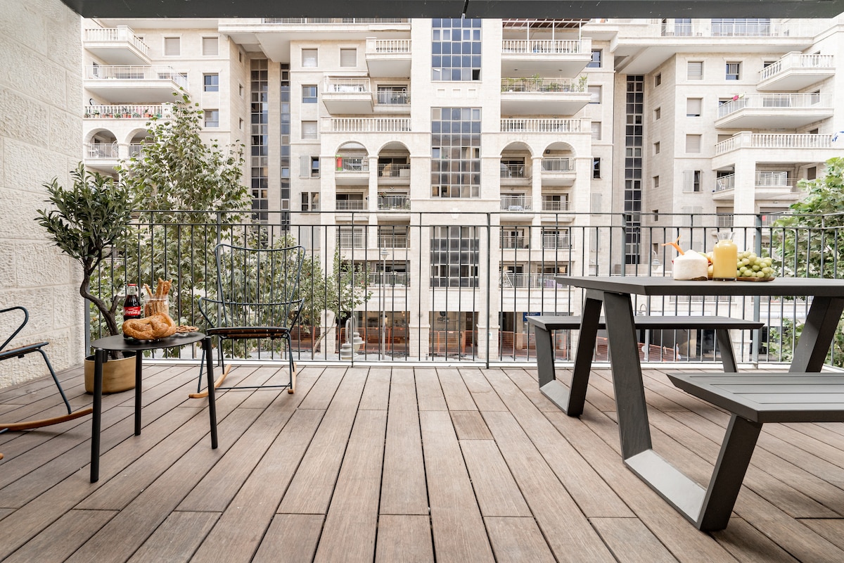 A spacious terrace features a dining table paired with benches, surrounded by wooden flooring. Several chairs are positioned nearby, and potted plants add a touch of greenery. The view includes adjacent apartment buildings, with large windows and balconies visible in the background.