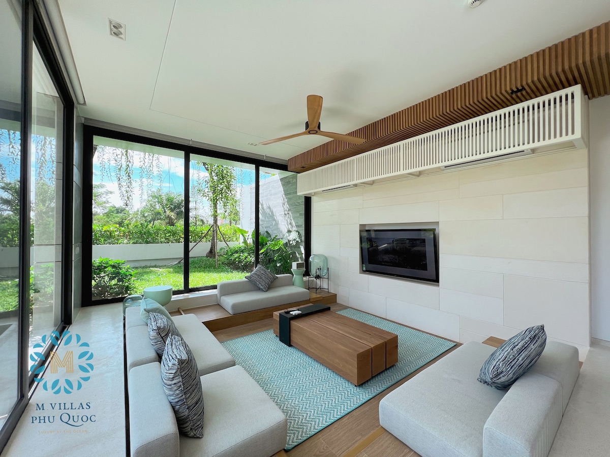 A spacious living room is showcased, featuring comfortable seating arrangements with light grey cushions. A wooden coffee table occupies the center, while large glass doors provide views of the green surroundings. A flat-screen television is mounted on the wall, enhancing the modern decor.