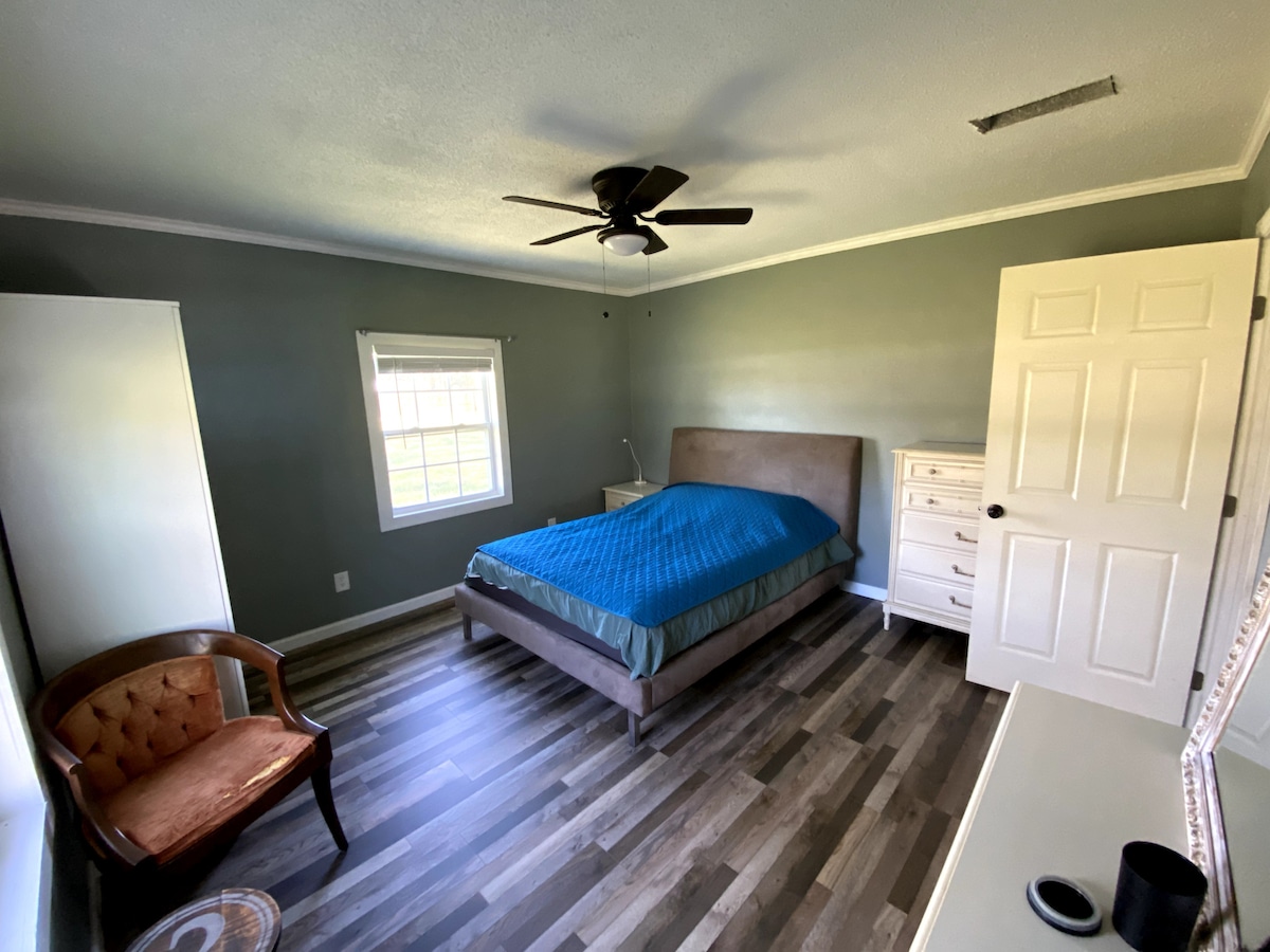 A spacious bedroom features a large bed covered with a blue blanket. A wooden chair adds a touch of warmth, and a dresser stands against one wall. Natural light enters through a window with a simple treatment, creating a calm and inviting environment.