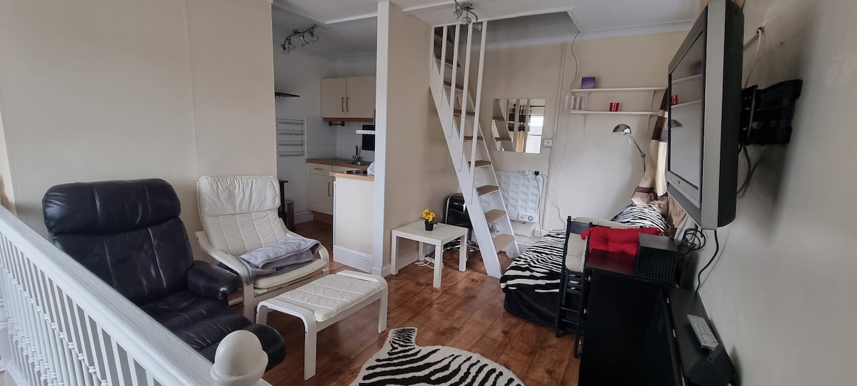The studio flat features a loft sleeping area and a cozy living space furnished with a black recliner, a white side table, and a patterned rug. A modern kitchen is visible in the background, complemented by natural light and neutral-toned walls.