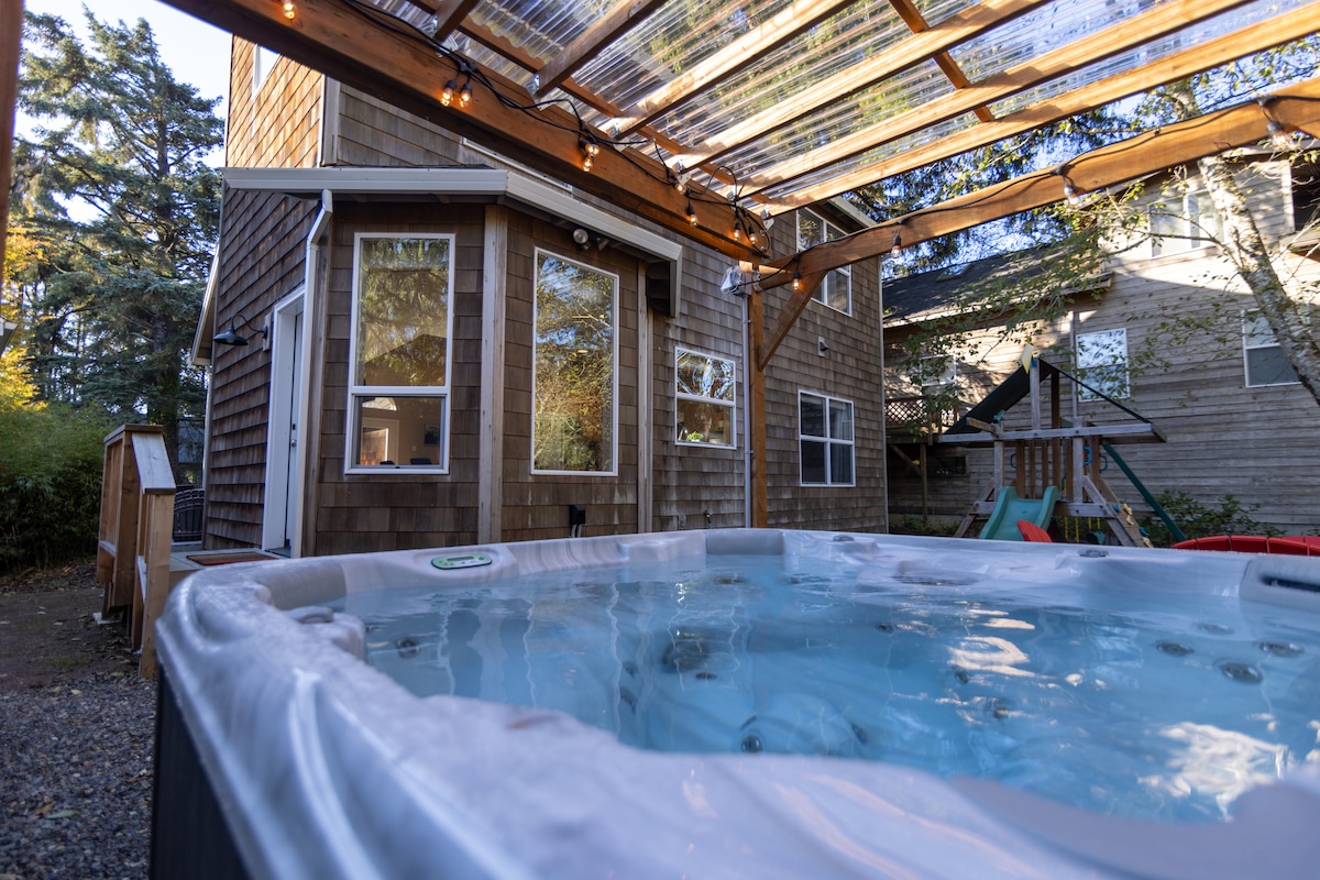 A spacious hot tub is positioned beneath a wooden pergola, surrounded by trees and the home's exterior. Large windows provide views into the living area. Colorful outdoor seating and a children’s playset are visible in the background, creating a relaxing outdoor retreat.