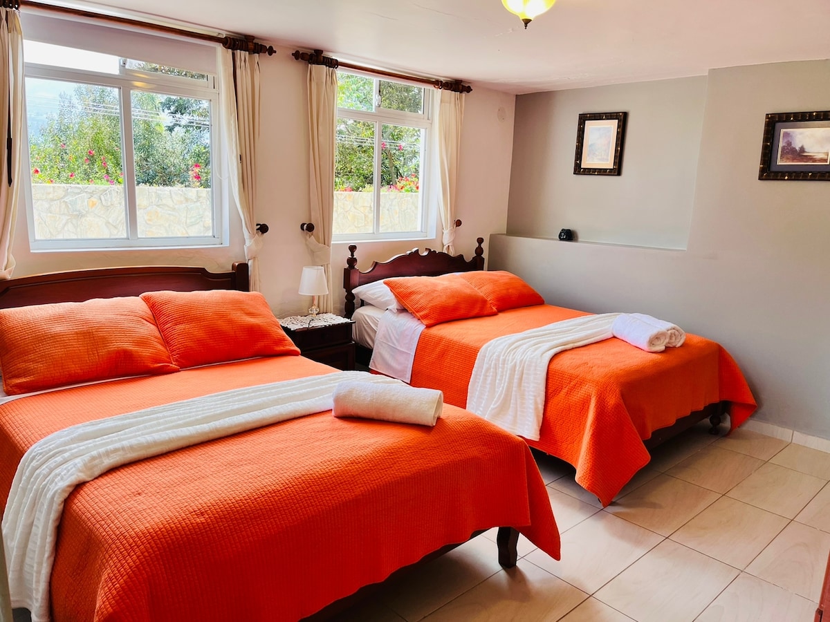 A comfortable sleeping area features two spacious beds dressed in orange covers, complemented by white linens. Natural light enters through large windows, showcasing garden views. Soft light fixtures and framed artworks add to the welcoming ambiance of the room.