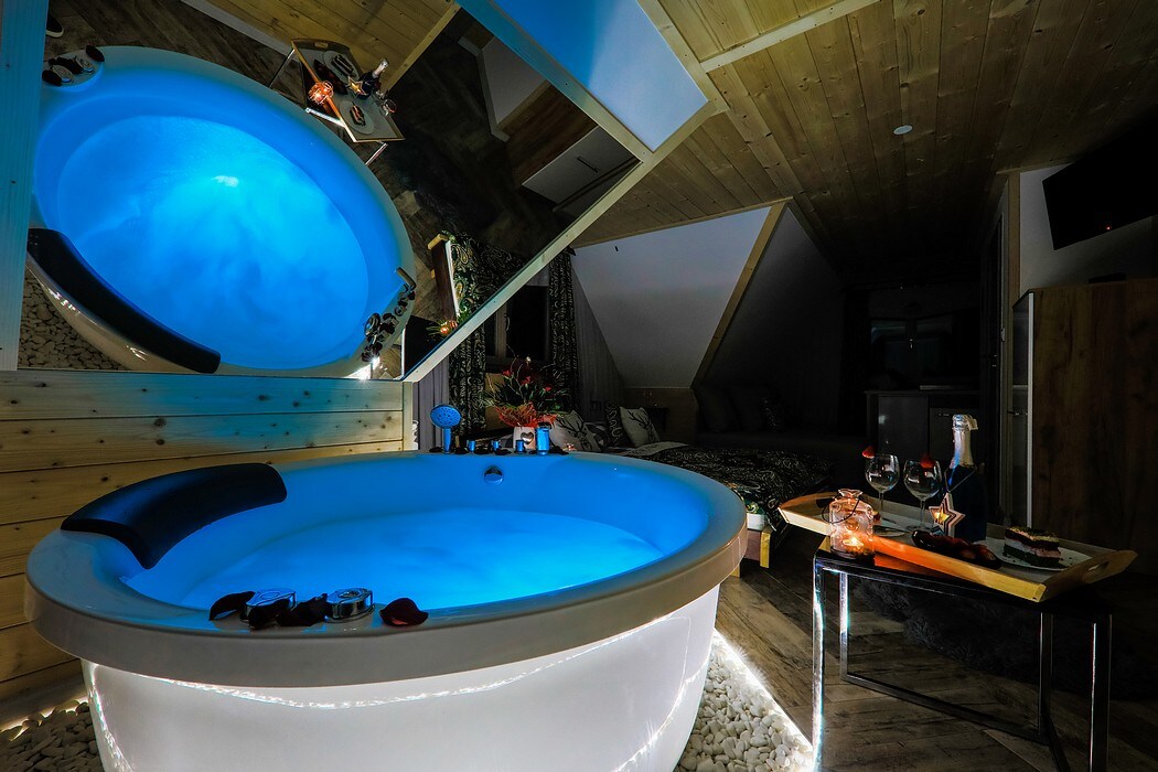 A spacious private jacuzzi is illuminated with soft blue lights, creating a calming atmosphere. Surrounding elements include wooden finishes and decorative items, enhancing the modern yet rustic appeal of the interior. A mirror reflects the jacuzzi, adding depth to the space.