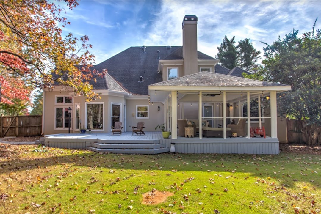 The exterior view features a spacious wooden deck surrounded by a well-maintained lawn. A screened-in porch invites relaxation, while large windows reveal the interior. A chimney rises from the roof, and autumn leaves scattered on the ground add a seasonal touch.