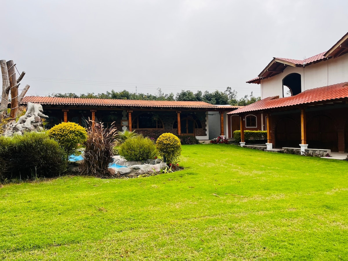A well-maintained garden area features neatly trimmed green grass and various shrubs. The building design includes a red-tiled roof and wooden accents. A stone water feature adds a tranquil element, while trees and plants provide a natural setting.