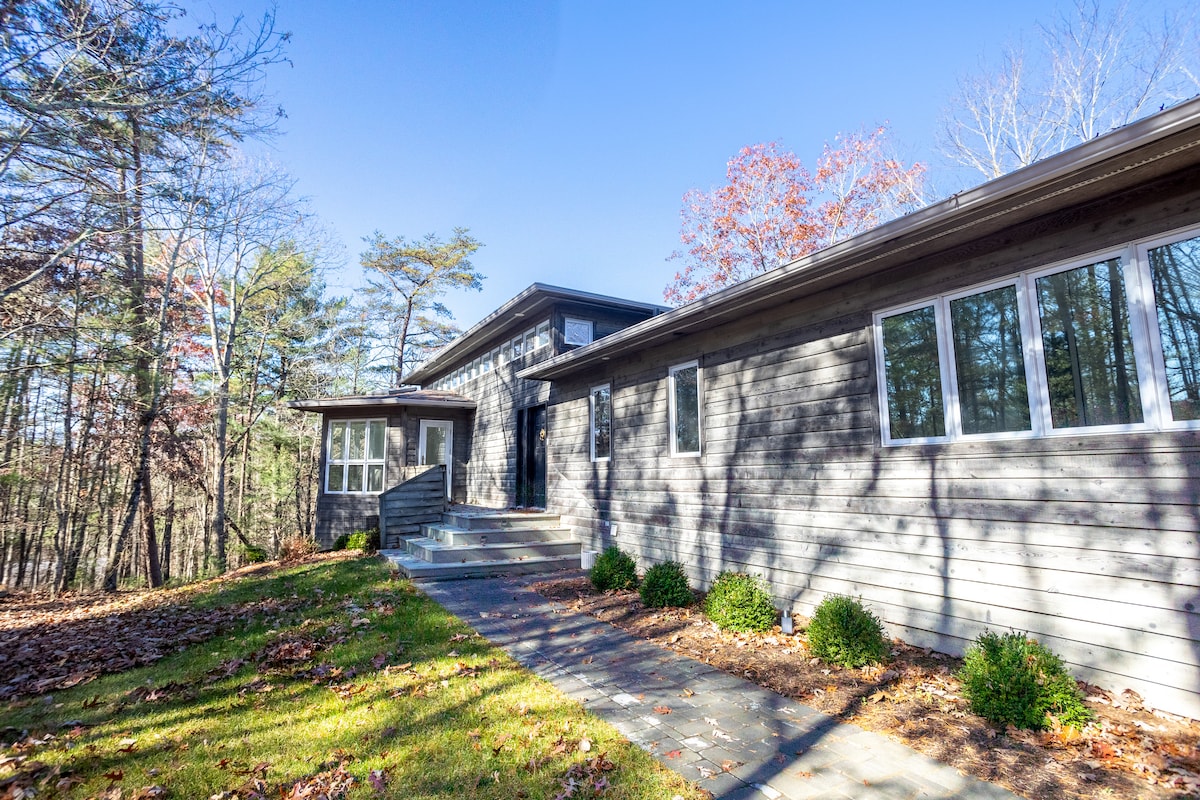 The modern exterior of the house features a combination of wood and large windows. A stone pathway leads to the front entrance, surrounded by well-maintained grass and shrubs. The surrounding trees provide a natural backdrop, showcasing the peaceful mountain setting.