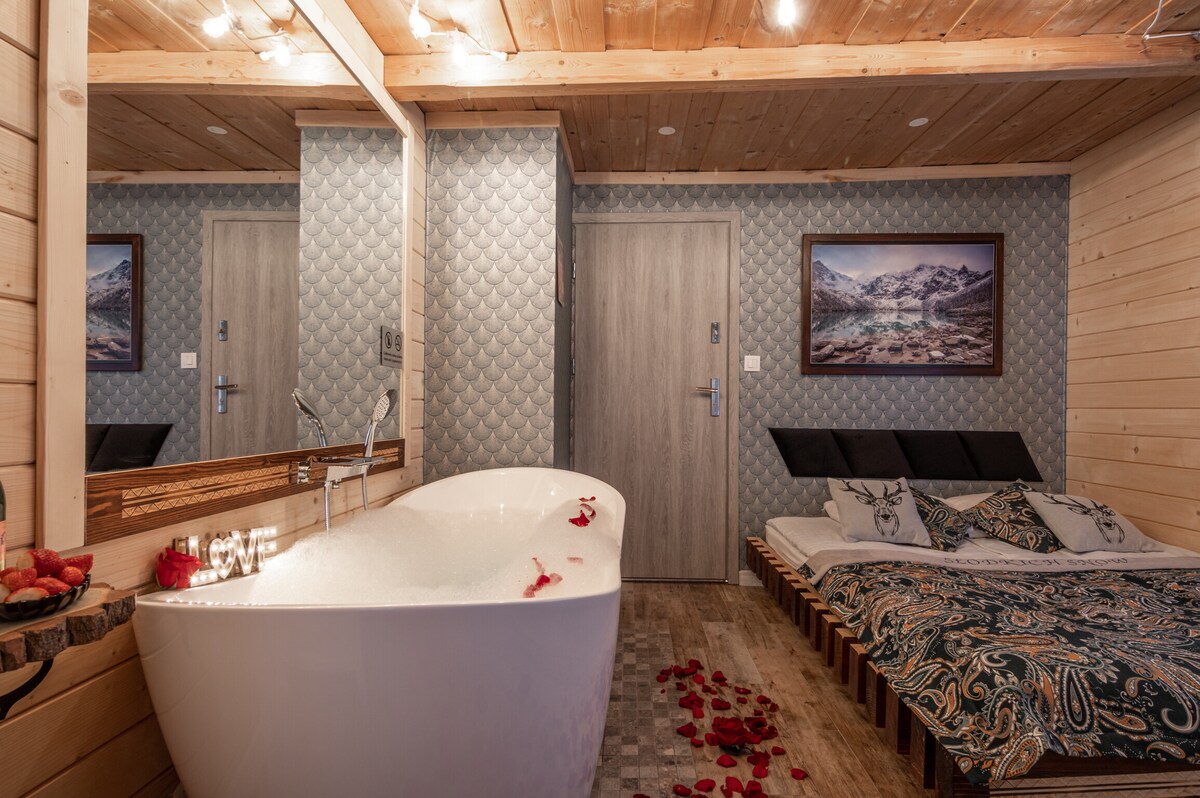 A modern room features a freestanding bathtub surrounded by rose petals, alongside a spacious bed with decorative bedding. Two mirrors enhance the light in the room, which showcases wooden paneling and cozy accents, creating a relaxing atmosphere.