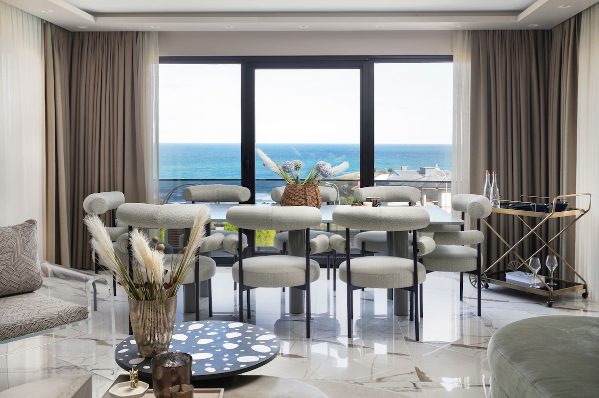 A bright dining area is presented with large windows offering views of the coastline. Circular dining tables are surrounded by upholstered chairs in neutral tones. Decorative elements include a textured vase filled with dried florals, complemented by a stylish bar cart in the corner.