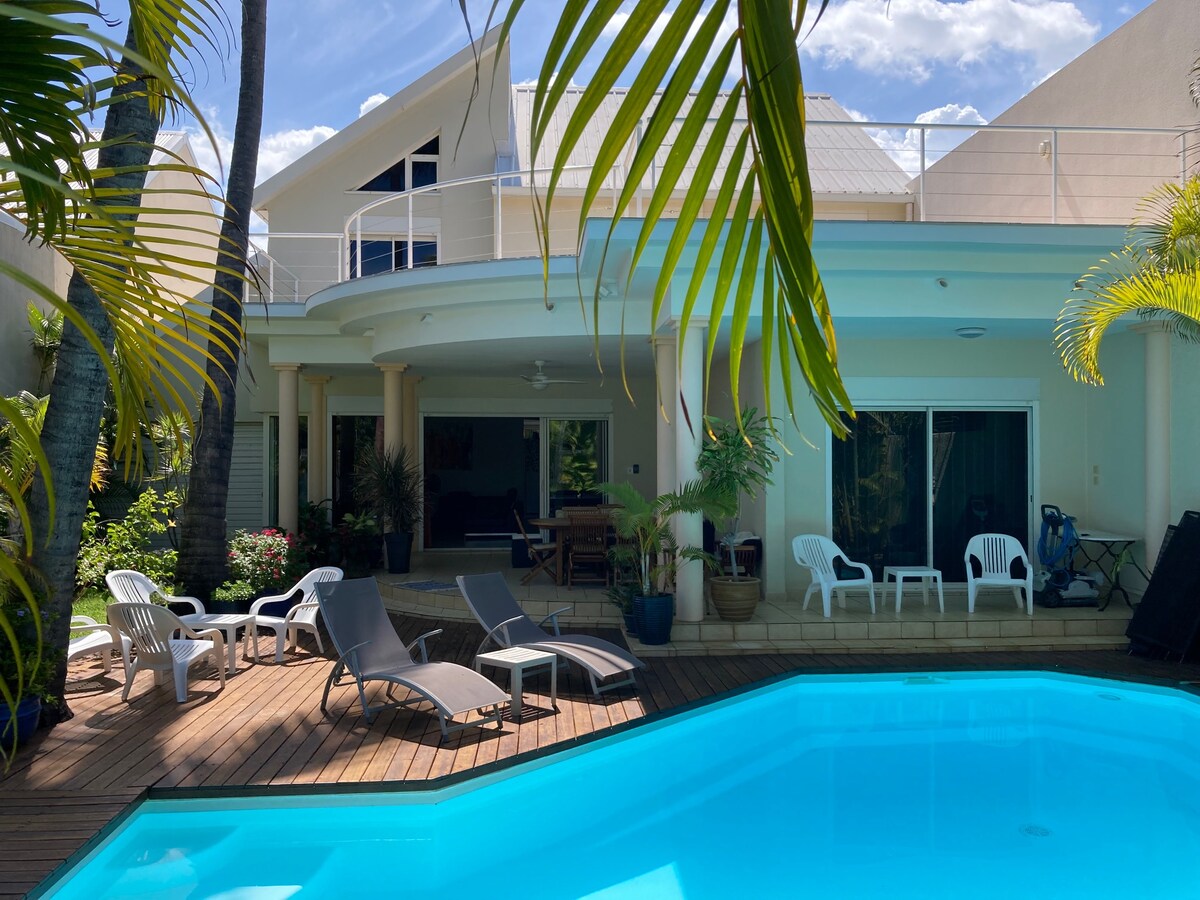 The outdoor area features a glistening pool surrounded by comfortable lounge chairs and shade from palm trees. The villa's spacious terrace, with sliding doors leading to the interior, offers a relaxing space for dining and lounging in a tranquil environment.