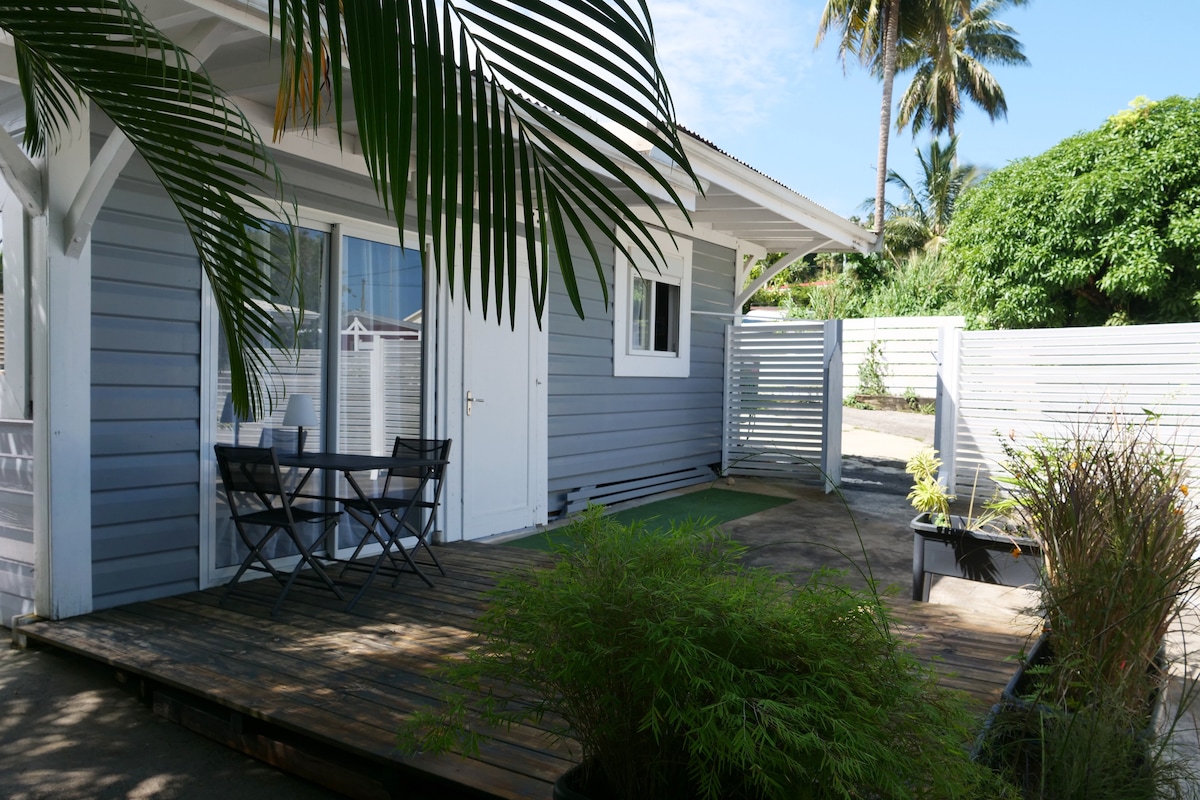 An outdoor deck area features a table with two chairs, surrounded by tropical greenery. The structure is clad in light gray wood with a clean white trim, showcasing doors that open to the interior. Nearby, a private, fenced area enhances the feeling of seclusion.