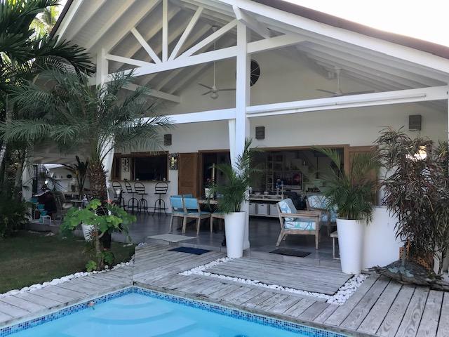 An inviting outdoor space is defined by a wooden deck surrounding a private pool, complemented by tropical plants. The open-plan structure features vaulted ceilings and large sliding doors that connect to a cozy living area adorned with comfortable seating.