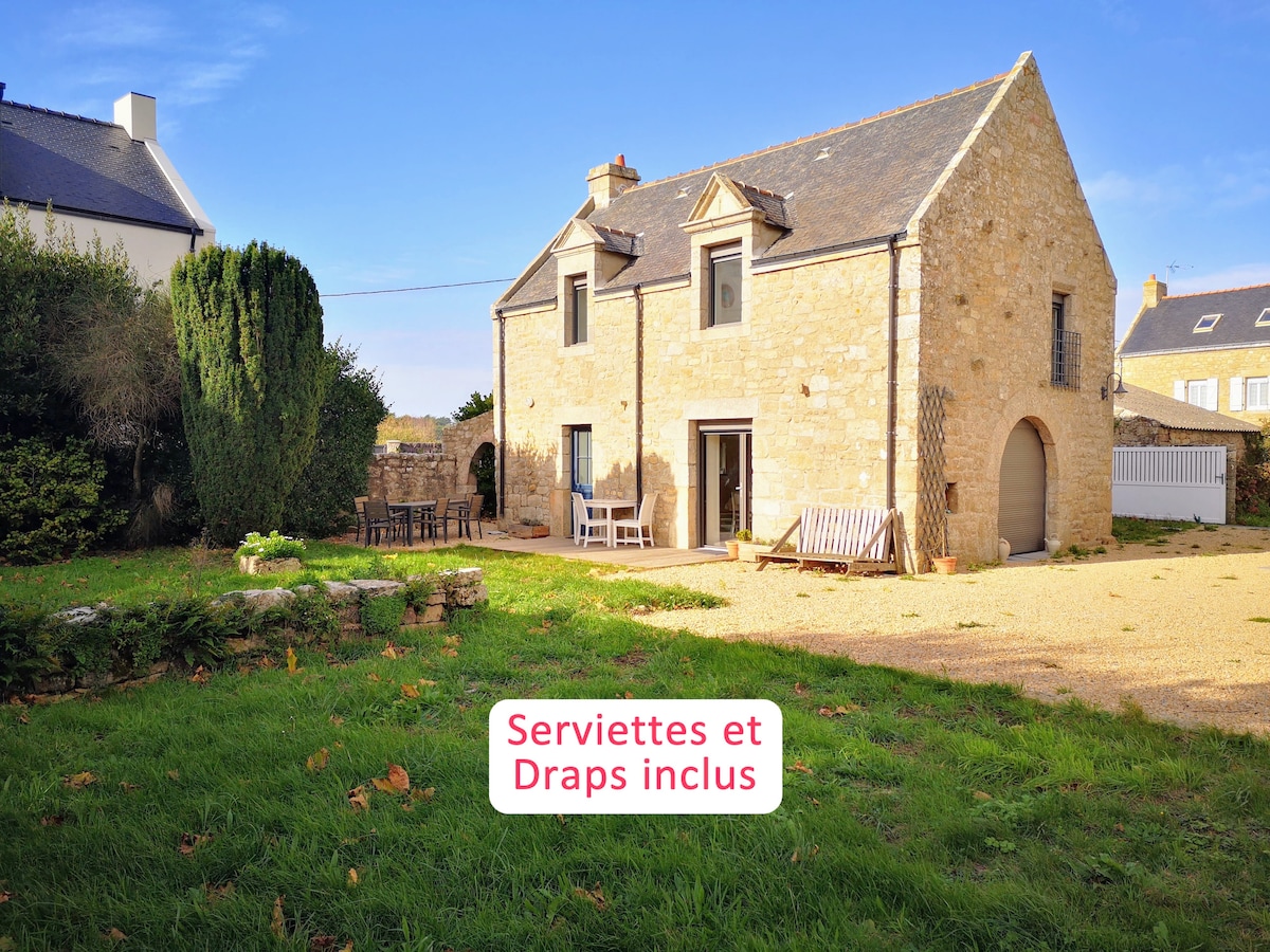 The stone house features a charming exterior with a mix of green grass and a gravel pathway leading to an outdoor seating area. The area is bordered by well-maintained shrubs and trees, enhancing the inviting outdoor space for relaxation.