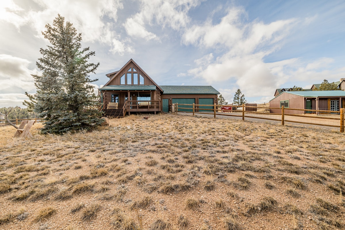 A spacious cabin is set against a backdrop of open terrain featuring a mix of grass and gravel. Large windows adorn the front, allowing natural light to fill the interior. Surrounding trees provide a natural boundary, and a wooden fence marks the property line.