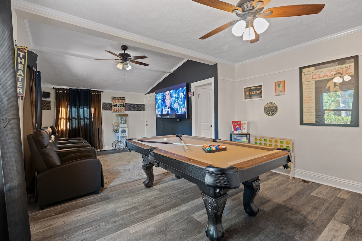 The game room features a classic pool table situated at its center, surrounded by comfortable seating for relaxation. A large screen is mounted on the wall for movie viewing, and poster art adds a nostalgic touch to the decor.