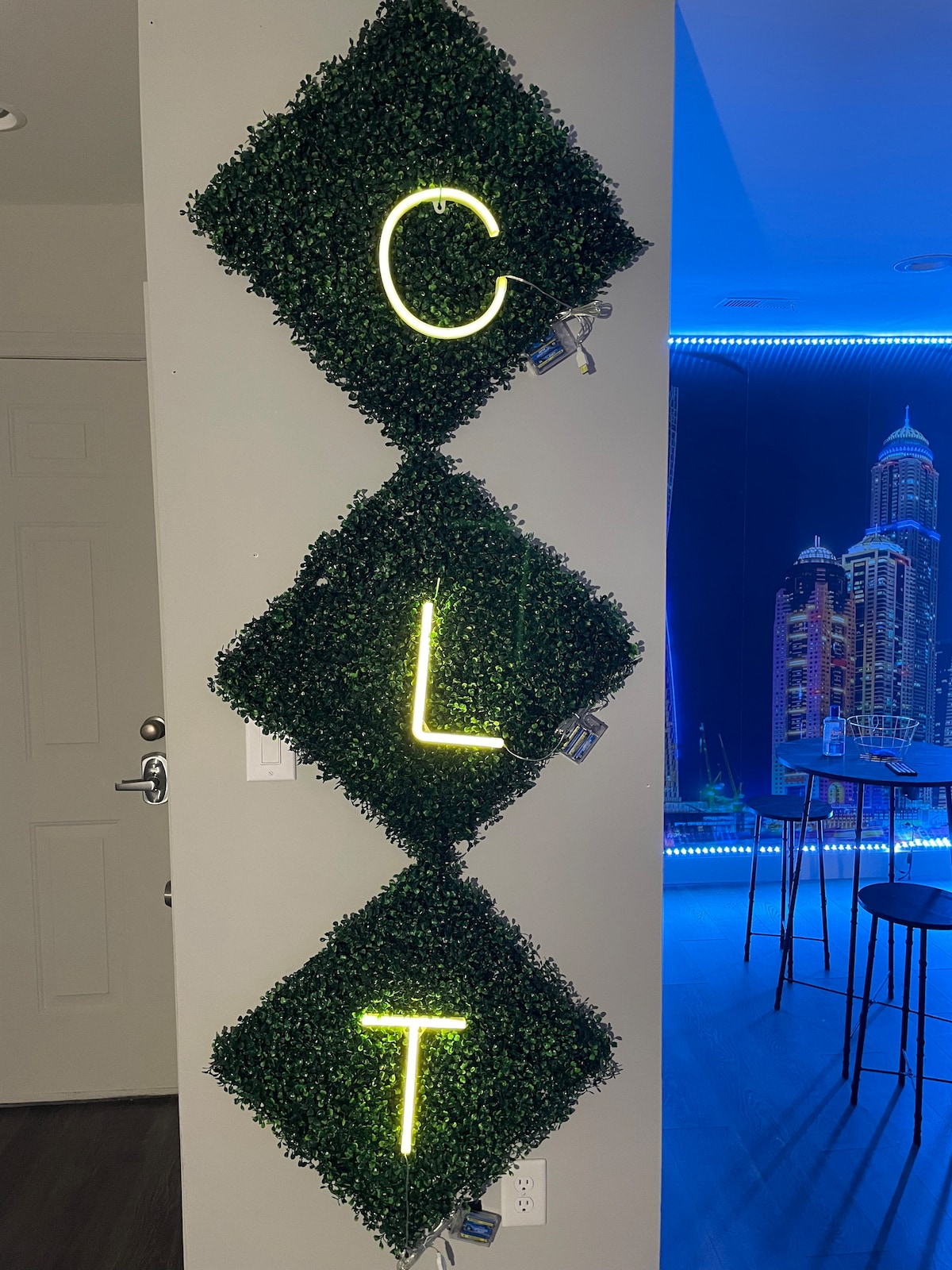 A wall feature showcases the letters 'C', 'L', and 'T' adorned in illuminated neon against a backdrop of artificial greenery. The setting is complemented by a modern cityscape view through a window, highlighting urban charm.