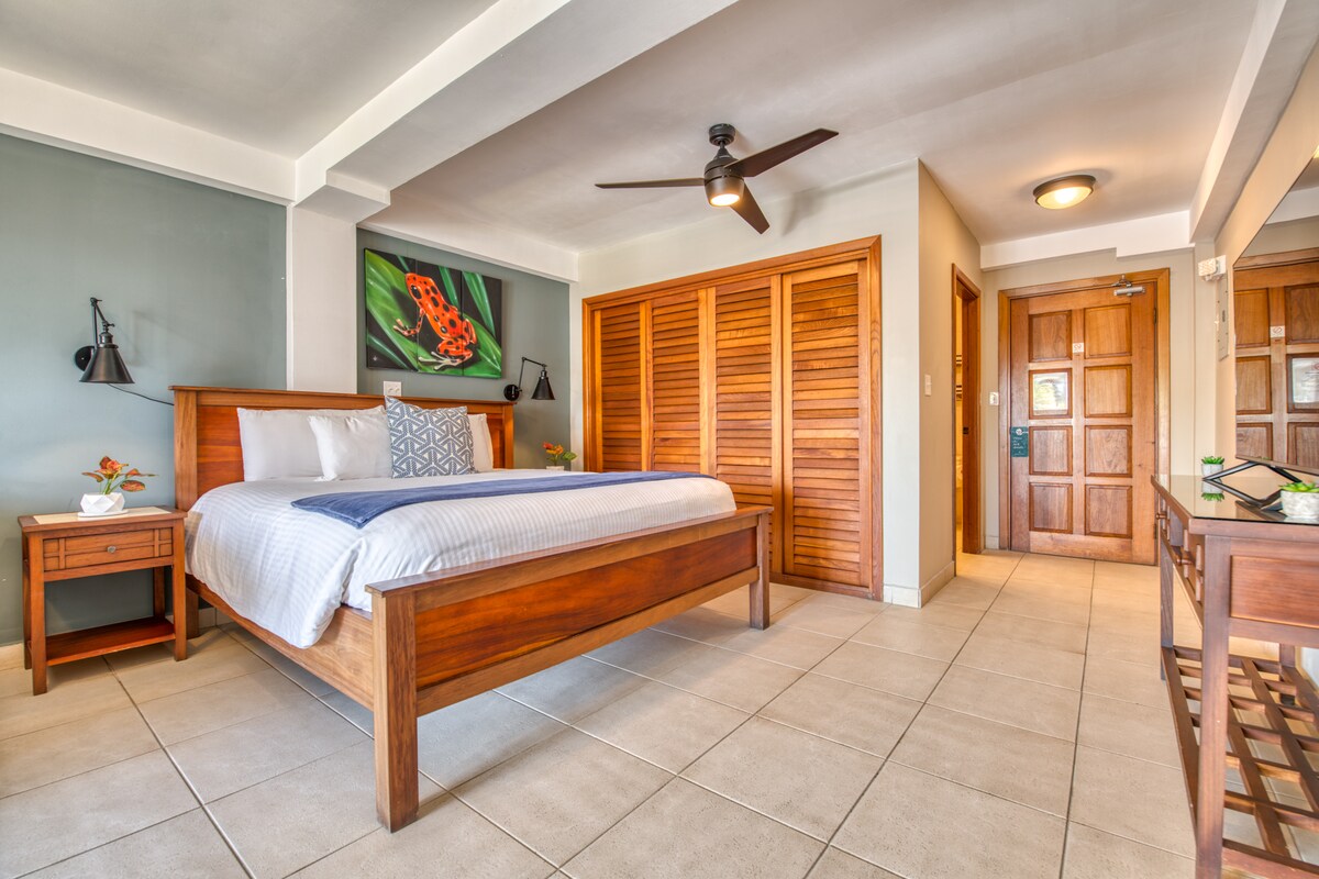 A bright and spacious sleeping area is furnished with a king-size bed, flanked by wooden nightstands. A large wooden wardrobe provides storage. Natural light fills the room from a nearby window. A ceiling fan is mounted, and artwork adds color to the walls.