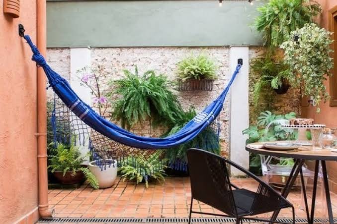A tranquil outdoor space features a blue hammock suspended between walls, surrounded by various lush green plants. A small dining table and a chair provide a cozy spot for relaxation, with textured tiles underfoot and soft lighting enhancing the serene atmosphere.