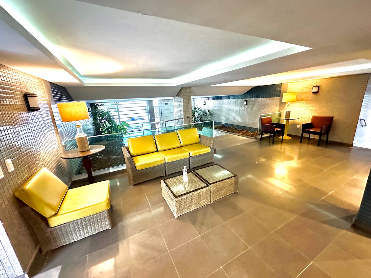 A modern lounge area is presented with comfortable yellow seating and a glass coffee table. Natural light filters through large windows, showcasing greenery in the background. Elegant light fixtures are placed strategically to enhance the space.