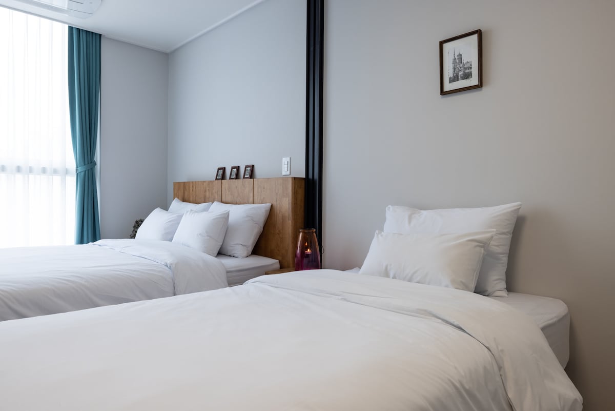A serene sleeping area is revealed, featuring two spacious beds with white bedding. Natural light filters through sheer curtains, illuminating the room. A wooden headboard adds warmth, complemented by decorative frames on the wall. A subtle lamp provides a soft glow beside one of the beds.