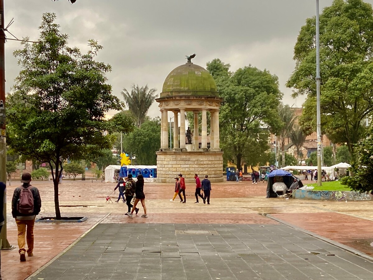 A plaza features a stone gazebo with a decorative dome, surrounded by green trees and people walking along the paths. A vibrant atmosphere is suggested by the presence of colorful structures in the background, while a gray sky looms overhead.