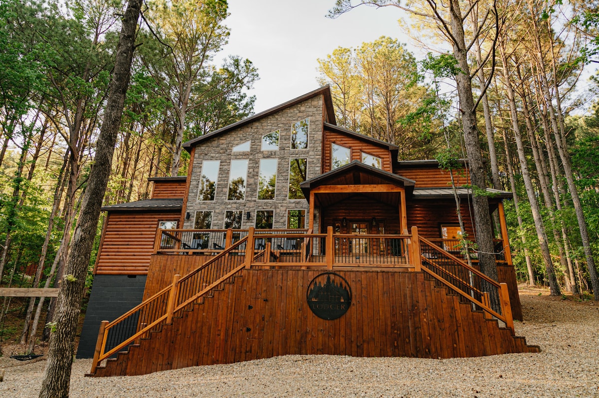 The cabin's exterior displays a mix of wood and stone, with large windows reflecting the surrounding tall pine trees. A grand entrance is highlighted by a spacious deck, featuring wooden railings that complement the natural setting. The cabin is positioned on a gently sloped hillside.