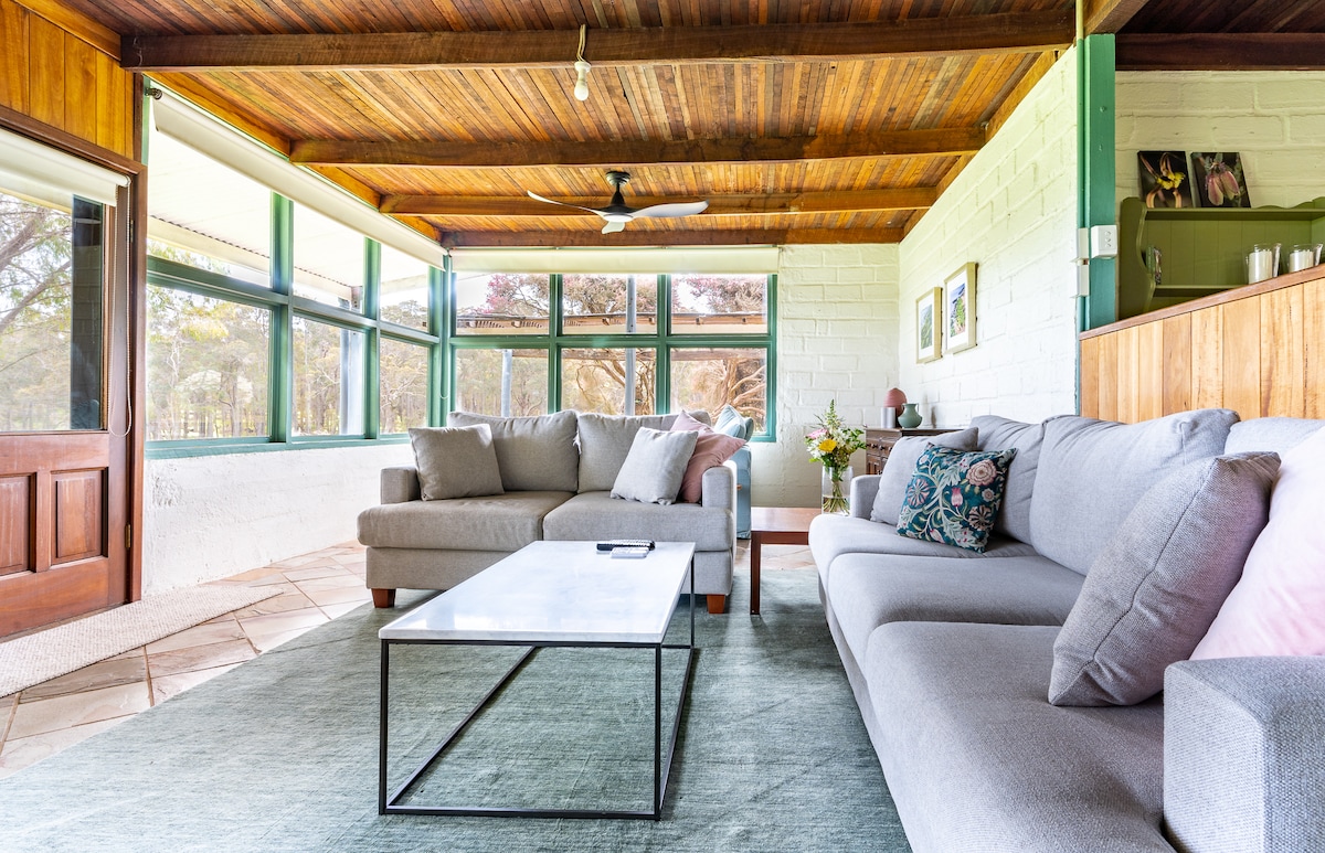 A comfortable living area features large glass windows that provide natural light and views of the surrounding nature. Two gray sofas provide seating, complemented by a low coffee table in the center on a soft area rug. The ceiling is adorned with wooden beams.