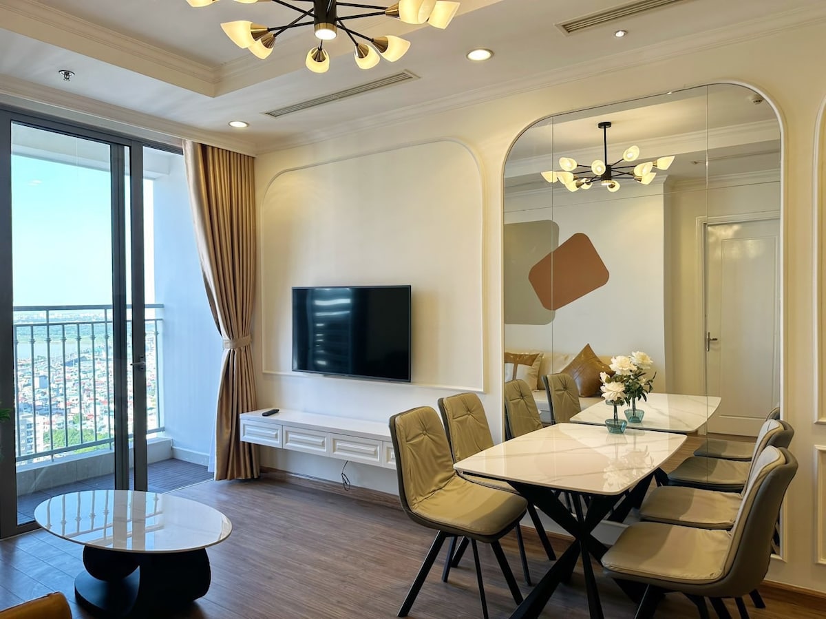 A modern dining area features a sleek table surrounded by six chairs. A television is mounted on the wall, opposite large windows offering a view of the city. Soft natural light illuminates the space, with decorative accents visible.