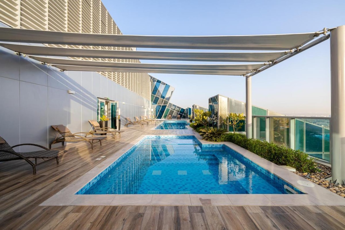 An outdoor pool is set on a rooftop terrace, surrounded by sleek lounge chairs and lush greenery. Sunlight reflects off the water's surface, highlighting the modern architecture of the building and providing an inviting space for relaxation.