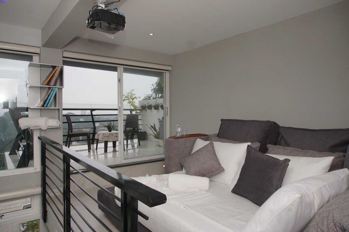 A modern studio room features a spacious sofa bed, positioned near large windows that showcase the outdoor balcony. A projector is mounted on the ceiling, and soft pillows adorn the sofa. The calming grey walls complement the serene atmosphere.