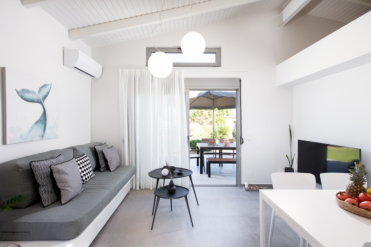 A modern living area features a grey sofa adorned with cushions and a minimalistic coffee table. Natural light enters through a sliding glass door, leading to an outdoor seating area under an awning. A flat-screen TV is positioned adjacent to a simple dining table.
