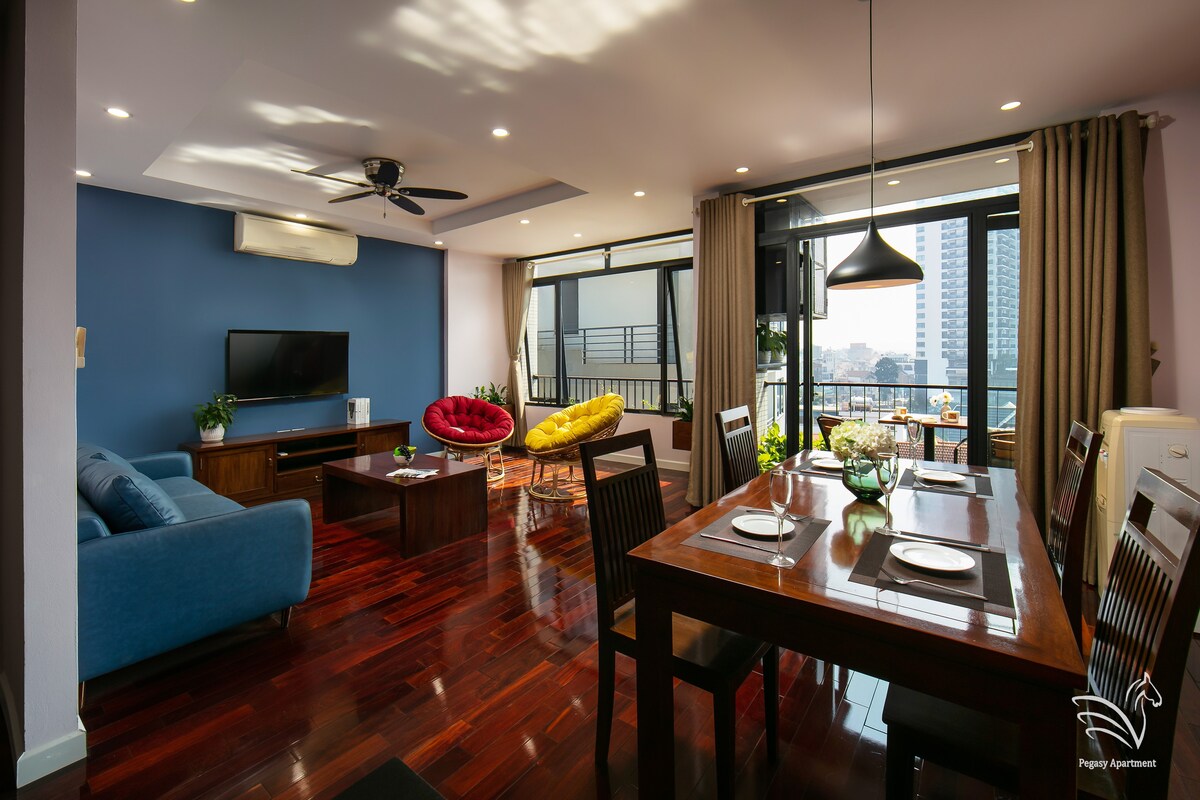 A bright living area is presented, featuring a comfortable blue sofa and a sleek wooden dining table set for four. A colorful armchair adds a pop of color, and large windows provide ample natural light and city views. The space is adorned with decorative plants.