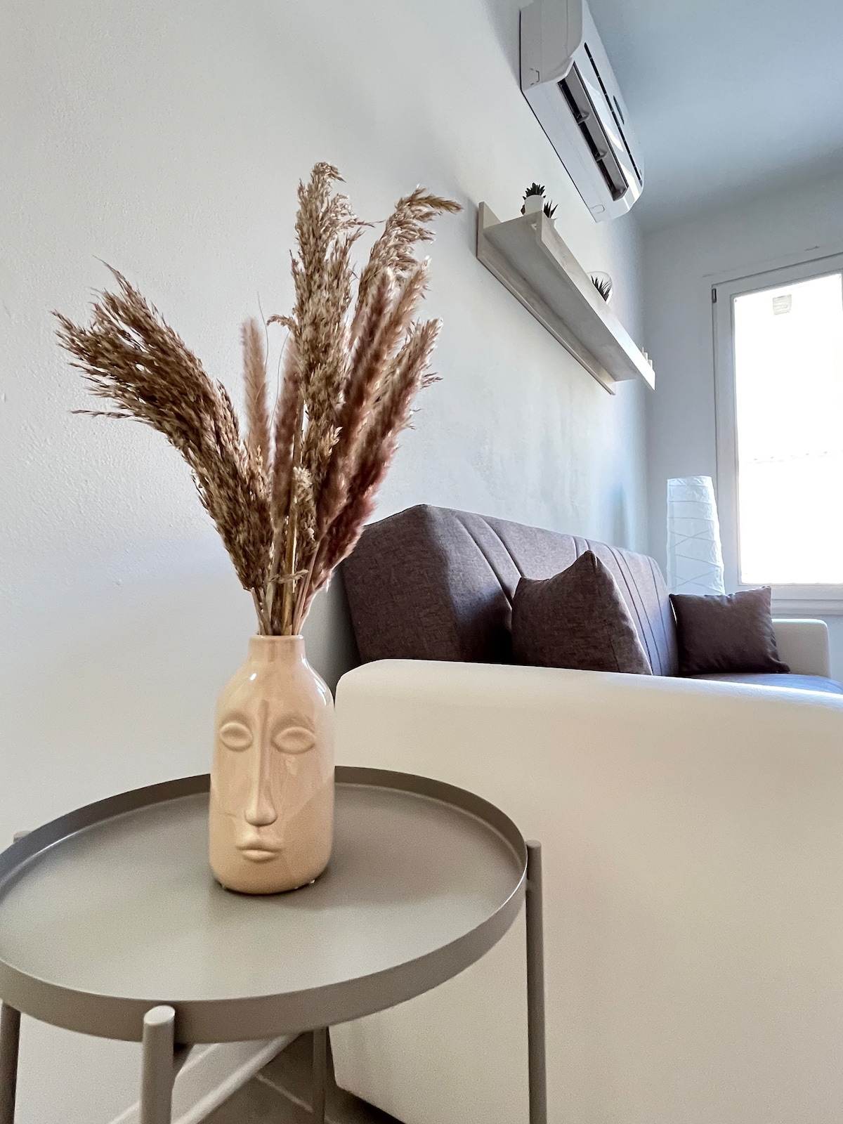 A side view of a soft, white sofa, accented by dark cushions, is highlighted near a sleek, glass table. Decorative dried flowers in a frosted vase add a touch of nature. A warm light from a nearby lamp and a wall-mounted air conditioning unit are visible.