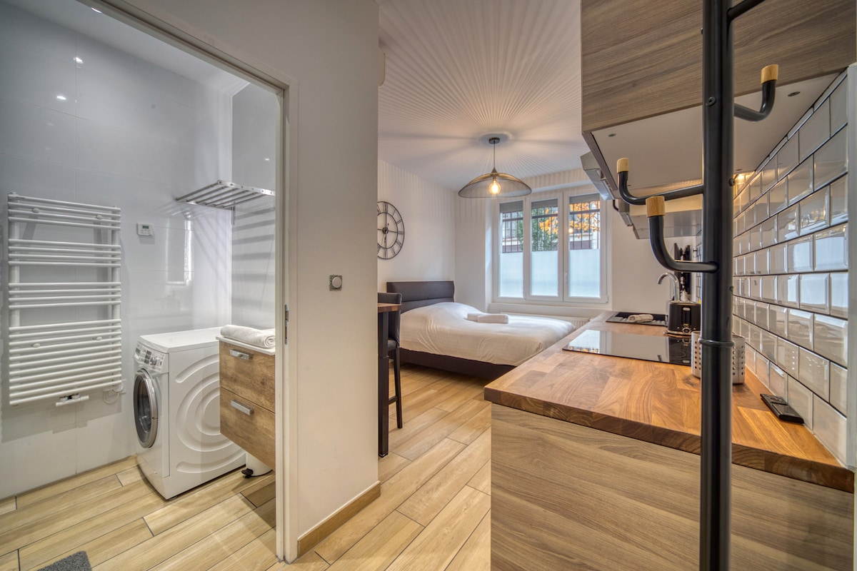 The image showcases a studio layout with an open design. A cozy bed is positioned near a large window, while a kitchen with modern appliances is visible on the right. A bathroom entrance with a washing machine and towel rack is located to the left.