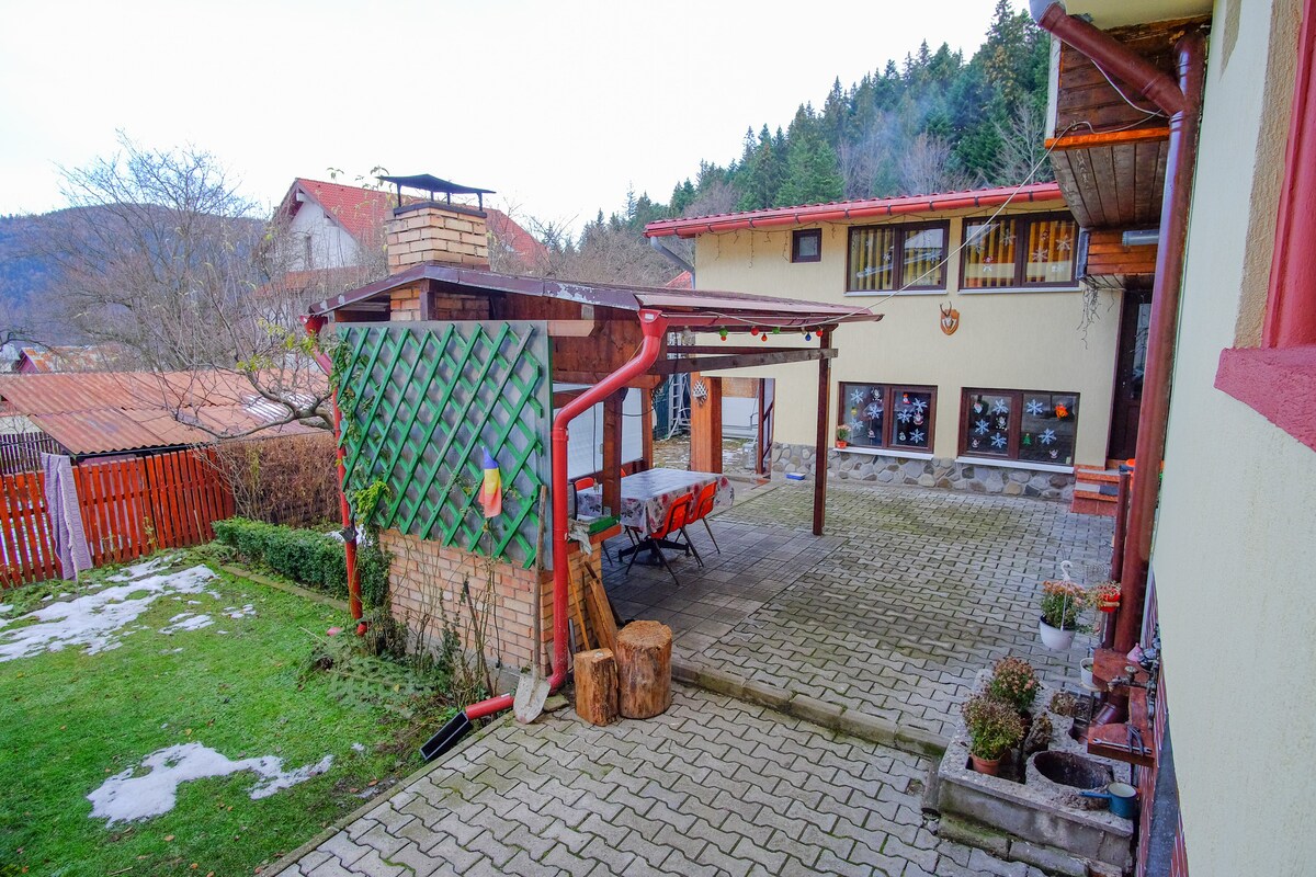 A spacious outdoor area features a stone patio with a covered seating area. Colorful chairs surround a large table, and greenery is present throughout the yard. A decorative trellis adds a natural element, while a peaceful backdrop of trees and distant hills is visible.