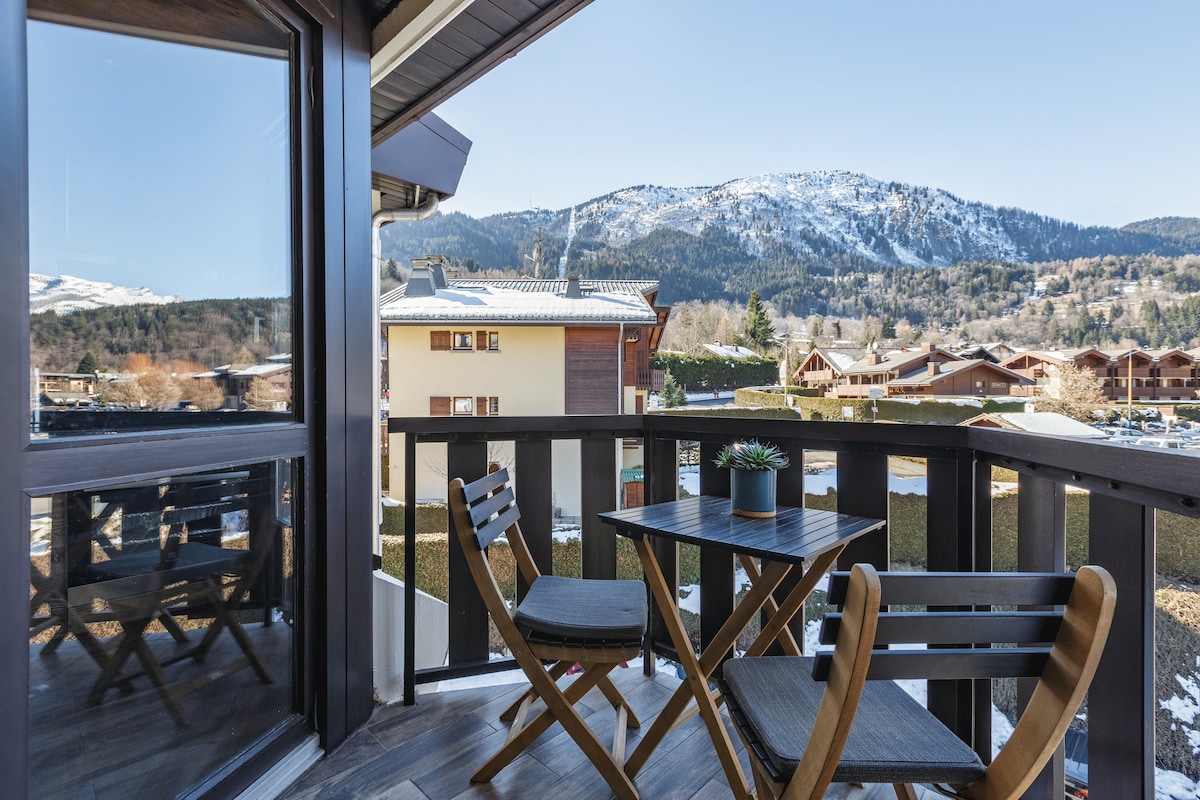A balcony view captures the surrounding mountains, providing a serene backdrop. The outdoor space is furnished with a small table and two folding chairs, inviting relaxation and enjoyment of the landscape.