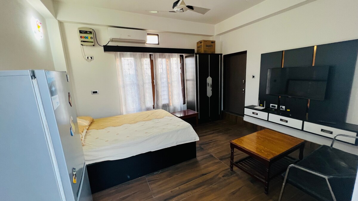 A modern bedroom features a spacious bed with a light-colored comforter. Large windows allow natural light to fill the room, partially covered by sheer curtains. A wooden coffee table and a comfortable chair are positioned beside a sleek entertainment unit.