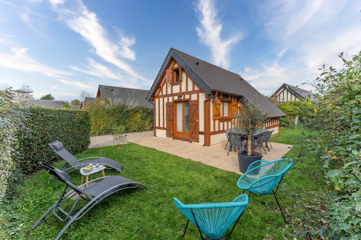 A charming chalet is surrounded by a landscaped garden, featuring a grassy area and a patio. Outdoor seating includes two loungers and a dining set with four chairs. The traditional facade displays wood accents and large windows, set against a backdrop of a blue sky with wispy clouds.