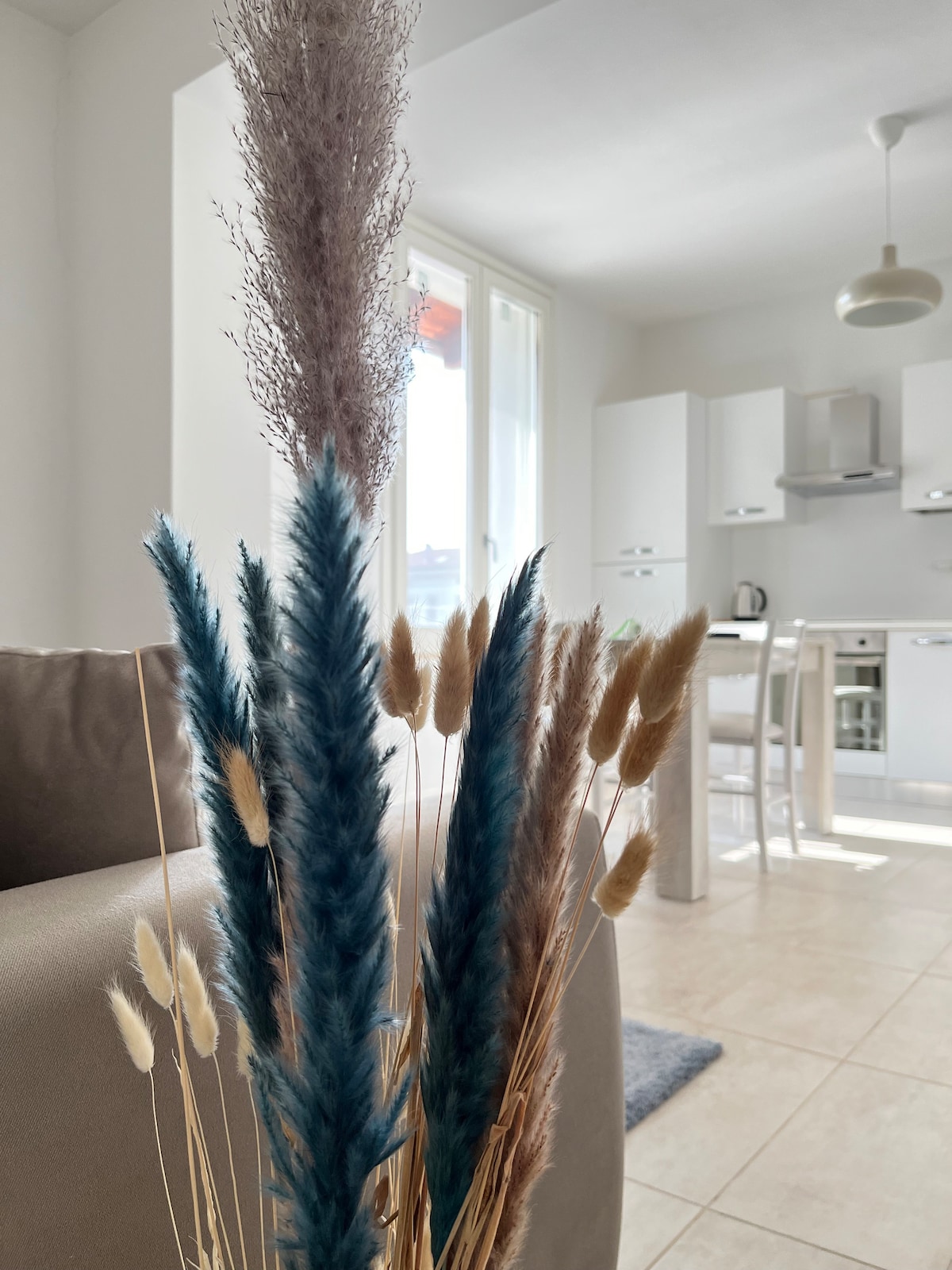 A decorative arrangement of dried plants is displayed prominently in the foreground. Soft blue and beige tones blend with natural textures. In the background, a bright kitchen area is visible, featuring white cabinetry and natural light filtering through large windows.