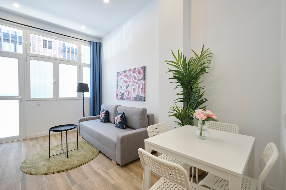 A bright living area features a gray sofa and a round black coffee table, surrounded by white dining chairs. A decorative plant and a floral artwork add a splash of color. Natural light streams through large windows, enhancing the overall openness of the space.