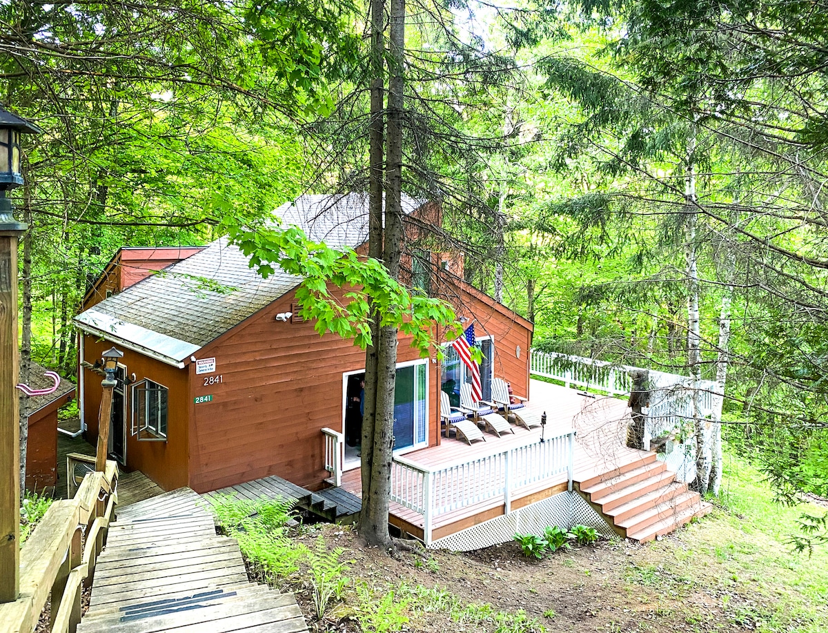 A charming ski chalet is nestled among the trees, featuring warm wooden siding and a large deck. Comfortable lounge chairs are positioned on the deck, which is accessible via a set of steps. Lush greenery surrounds the property, providing a serene and natural setting.