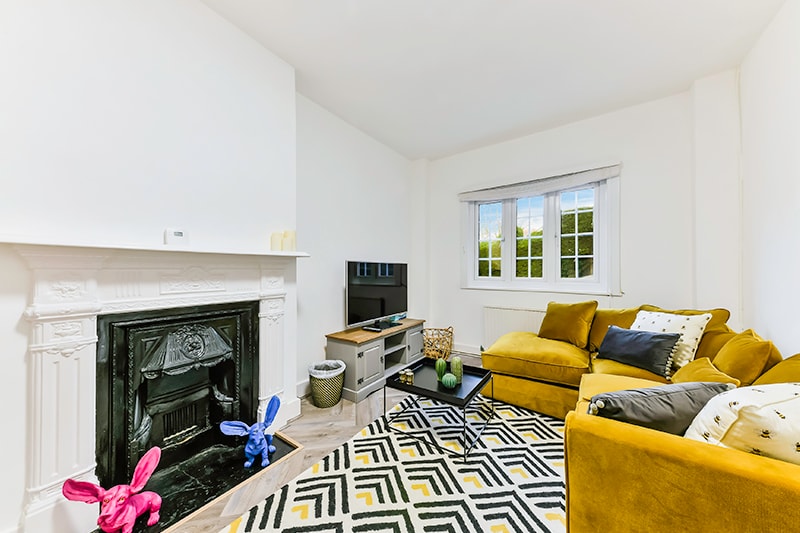 A cozy living area features a yellow sectional sofa and a patterned area rug. A fireplace is framed with white detailing, while a television is placed on a modern stand. Natural light enters through the window, illuminating the vibrant decor elements.