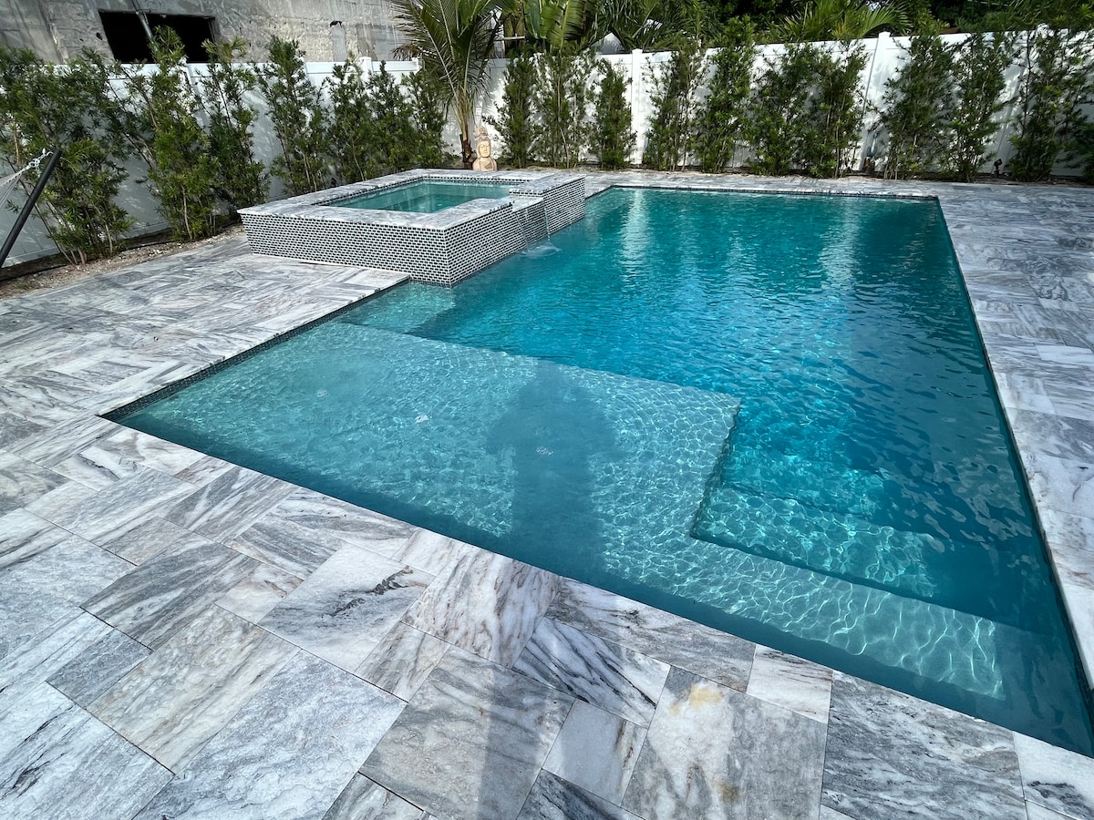 A clear view of a serene pool area features a spacious saltwater pool and a separate jacuzzi, both surrounded by elegant marble tiles. Lush green landscaping provides privacy along the perimeter, enhancing the tranquil setting.