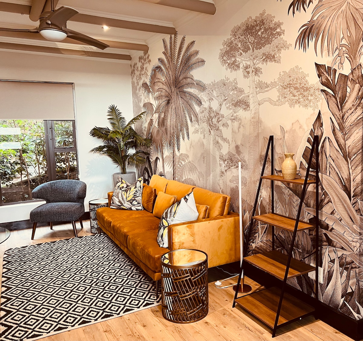 A vibrant lounge area features a mustard-colored sofa against a botanical mural. A patterned area rug adds texture to the floor, while an accent chair and an open bookshelf provide functionality. Large windows allow natural light to brighten the space, surrounded by greenery.
