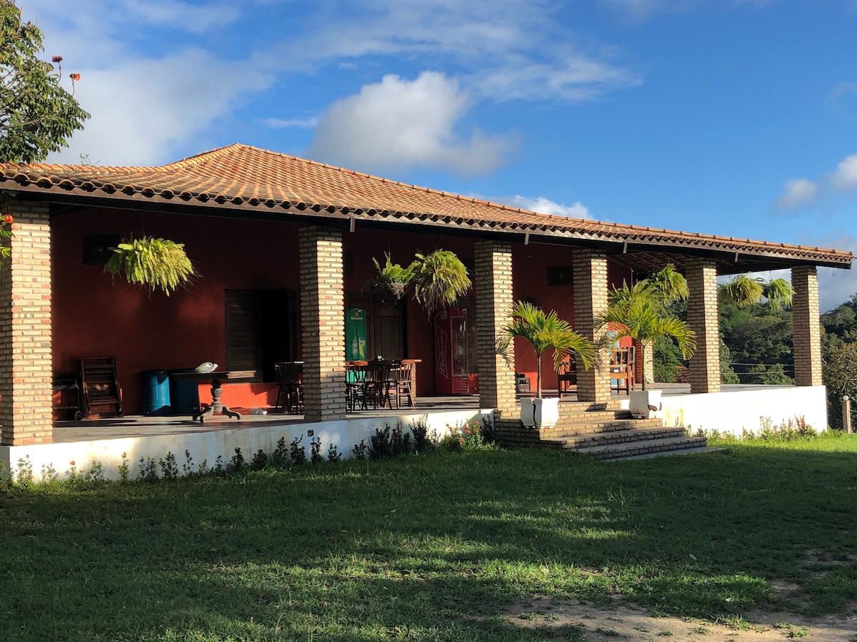 A single-story structure with a tiled roof is depicted, featuring a wide porch supported by brick columns. Lush greenery surrounds the area, and potted plants enhance the natural setting. Outdoor seating arrangements are visible, inviting relaxation and gatherings.