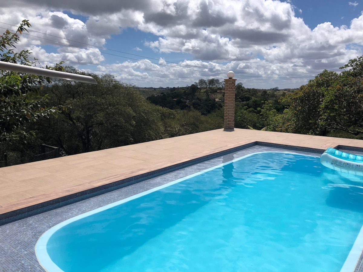 A clear swimming pool is positioned on a spacious deck, offering views of the surrounding greenery and distant hills under a partly cloudy sky. The pool features a gentle curve and a shallow area, inviting relaxation and enjoyment.