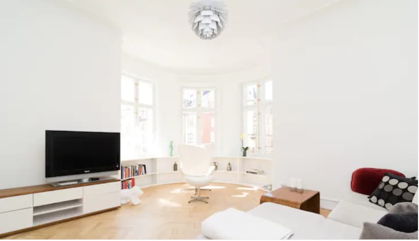 A spacious living room features a large window setup that allows natural light to fill the space. A television is positioned against a simple entertainment unit, and a comfortable chair is placed in the center. Light wood flooring contributes to a clean and airy ambiance.