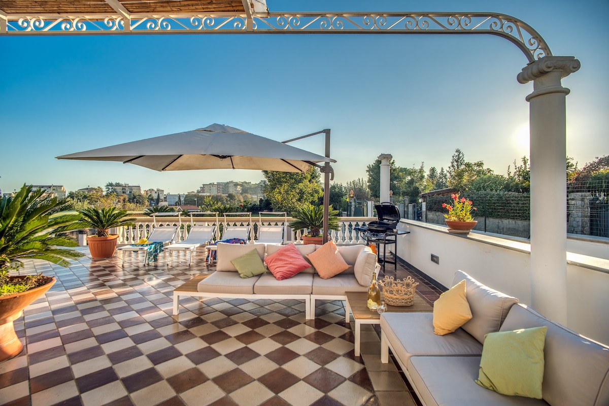A spacious terrace is highlighted by comfortable seating, accent pillows, and a large umbrella for shade. Potted plants add greenery, while a grill is positioned for outdoor cooking. The scene is complemented by a clear sky and soft sunlight casting gentle shadows.