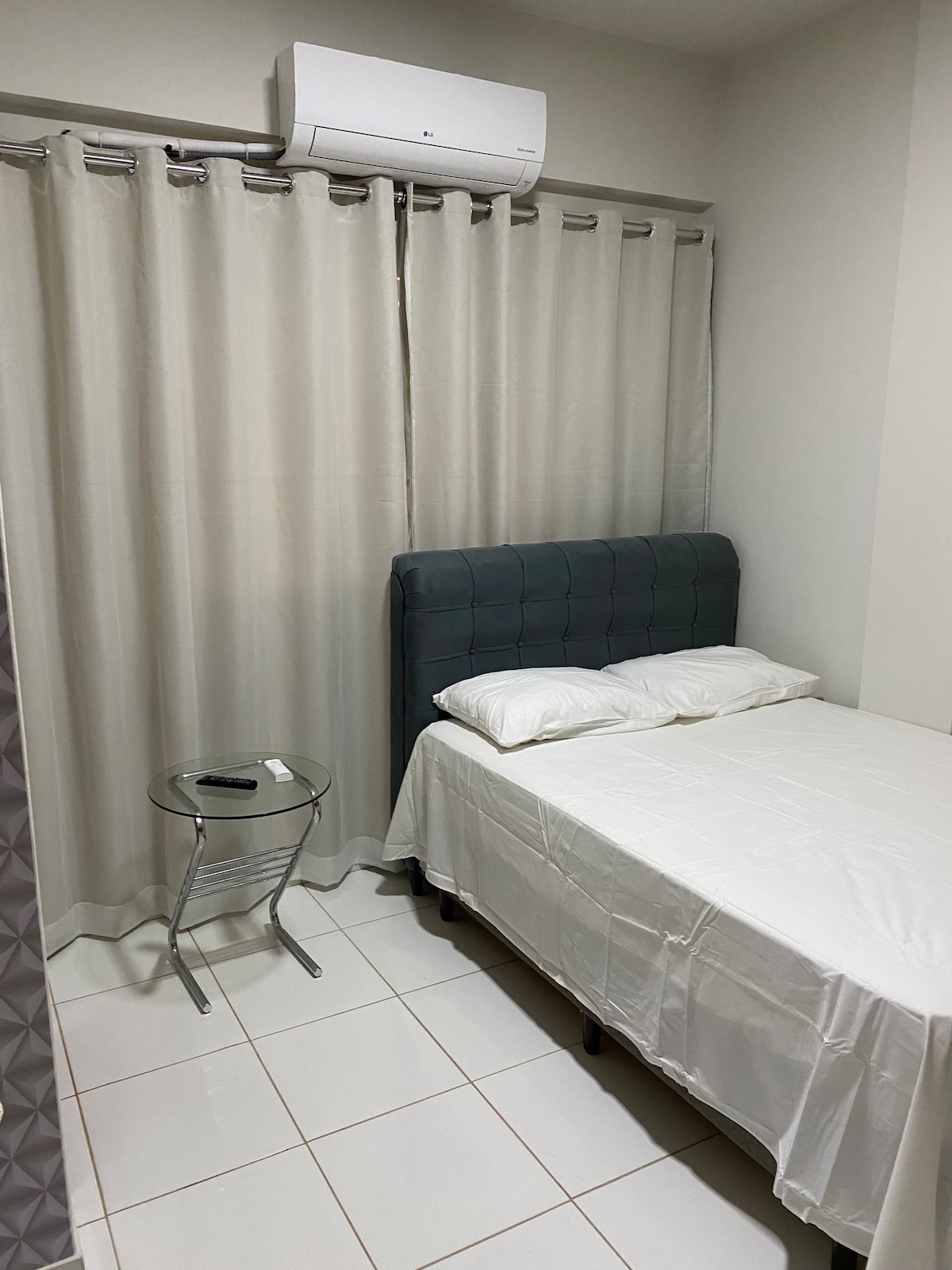 A simple bedroom features a spacious bed with white bedding against a muted wall. A small, round side table is positioned next to the bed. Light-colored curtains frame a window, and an air conditioning unit is mounted above for comfort.